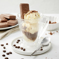 Glass mug with ice cream and chocolate sauce, surrounded by cookies and coffee beans on a white surface.