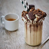 Chocolate milkshake with whipped cream and cookie pieces in a glass, next to a white mug on a wooden surface.
